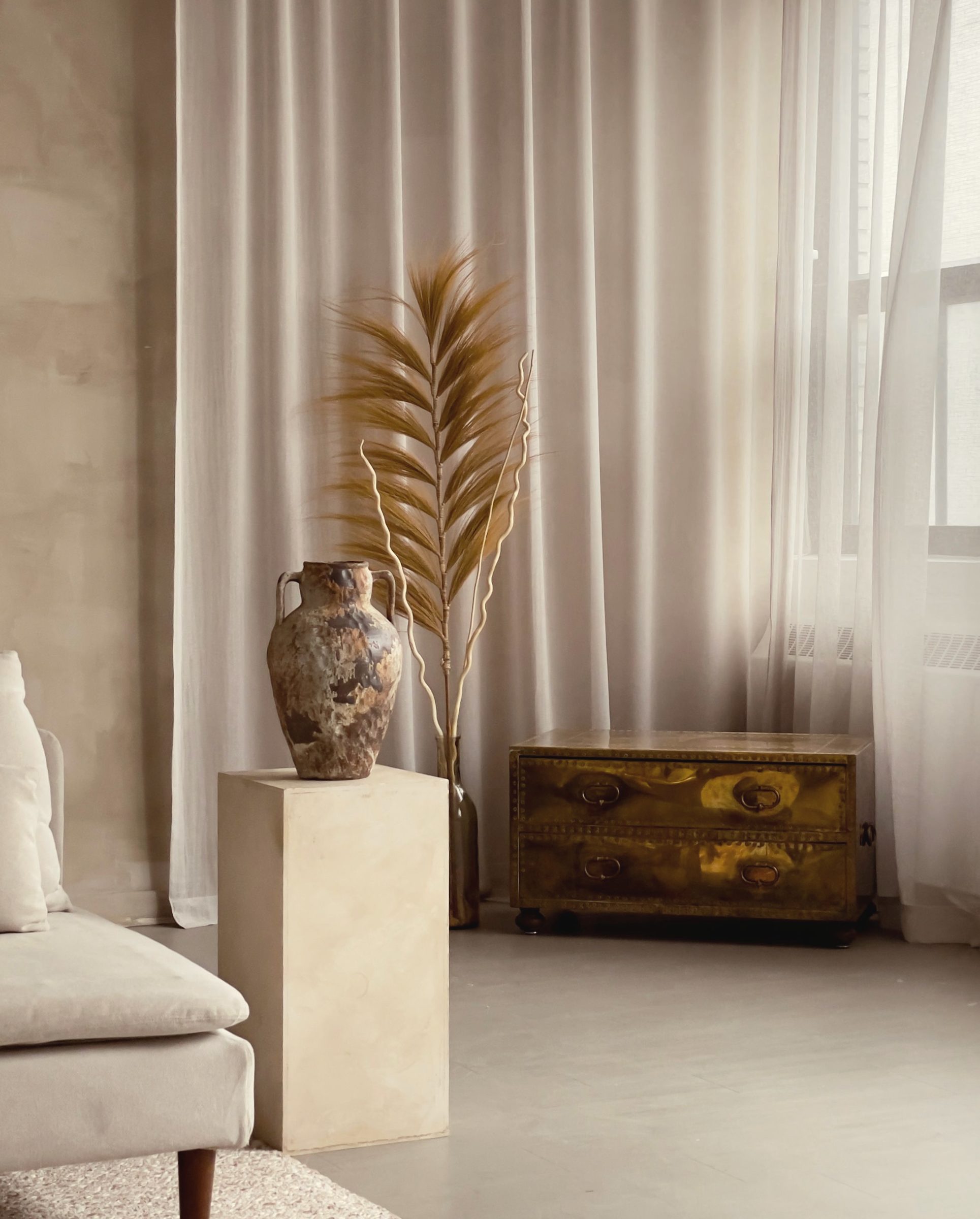 Sitting room with lime plaster walls, linen curtains, and a vessel of dried palm on a stone plinth