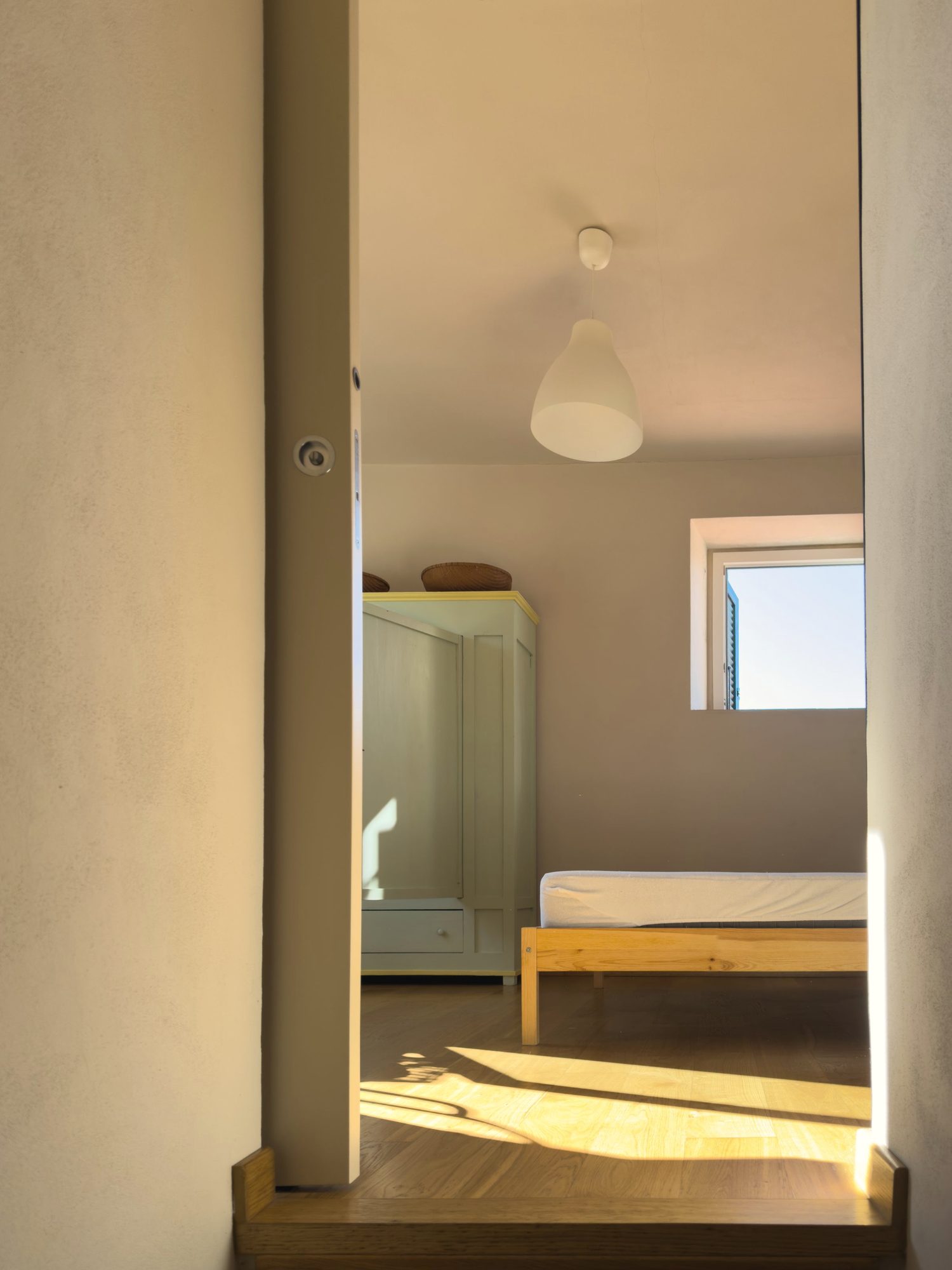 View through a doorway into a guest room with a pale pine bed in afternoon sun